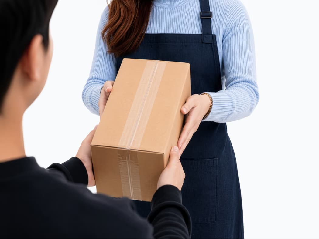 Woman in blue sweater and apron handing a taped cardboard box to a person in black.