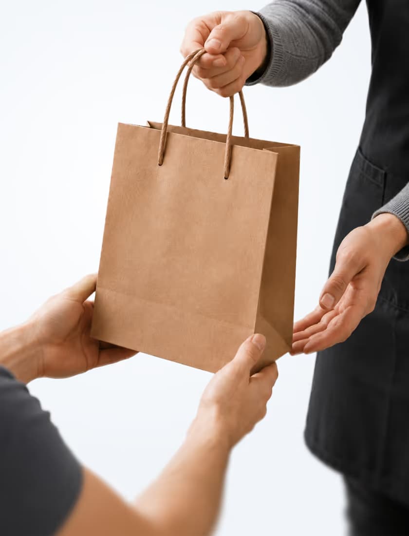 Person handing over a brown paper shopping bag to another person wearing a gray sleeve and black apron.
